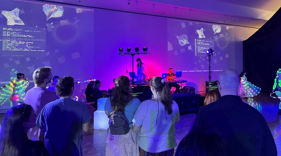 A wide shot of a large room washed in purple light. A crowd stands in the foreground facing a stage where two performers work on laptops. Behind them, large projections on the wall display lines of computer code alongside floating abstract 3D shapes. To the sides, dancers wearing LED-lit costumes perform.