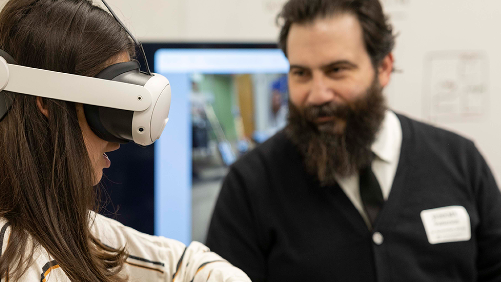 A woman stands in the foreground wearing a VR headset while a man helping to demo a project stands in the background, directing her.