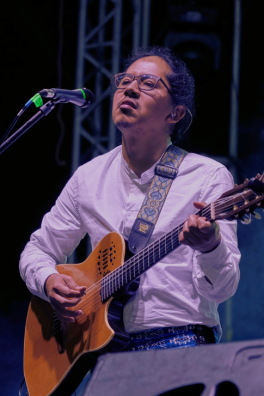 An action shot of Ati playing the guitar while standing on stage during a performance and singing into a microphone.