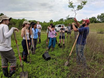 A group of students stand in a field with shovels while a man standing in the front, explaining how to plant.