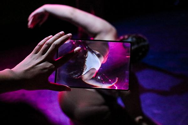 A hand holds a small plexiglass frame in front of a dancer who is spiraling their body. The frame flips and distorts the dancers image