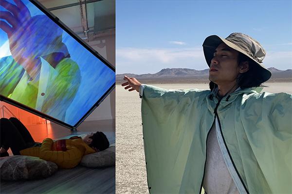 A man stretches his arms out in a rain parka and a sun hat with eyes close in a desert-like environment