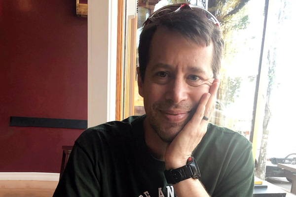 A photo of Peter Stuart, a man with short brown hair wearing a green t-shirt, and resting his head on his hand while smiling at the camera.