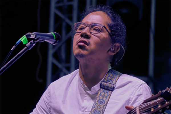 A headshot photo of Ati Cachimuel wearing a white shirt and glasses and singing into a microphone while playing the guitar.