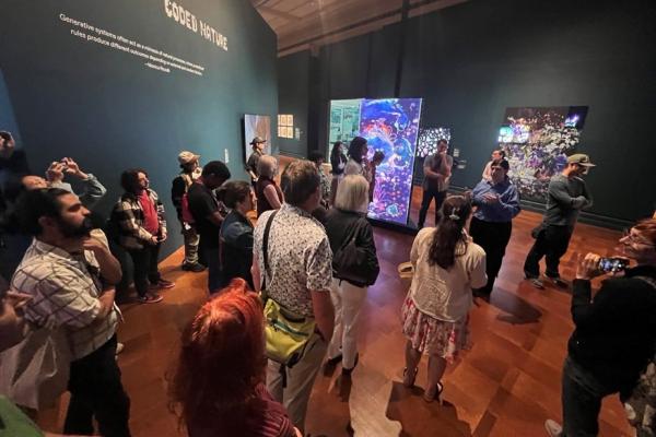 A group of approximately twenty people stand in a museum gallery with dark teal walls. They are attentively listening to a curator in a blue shirt who is speaking and gesturing. Behind the group, wall text reads "CODED NATURE," and several large, bright digital art screens display colorful, abstract floral imagery.